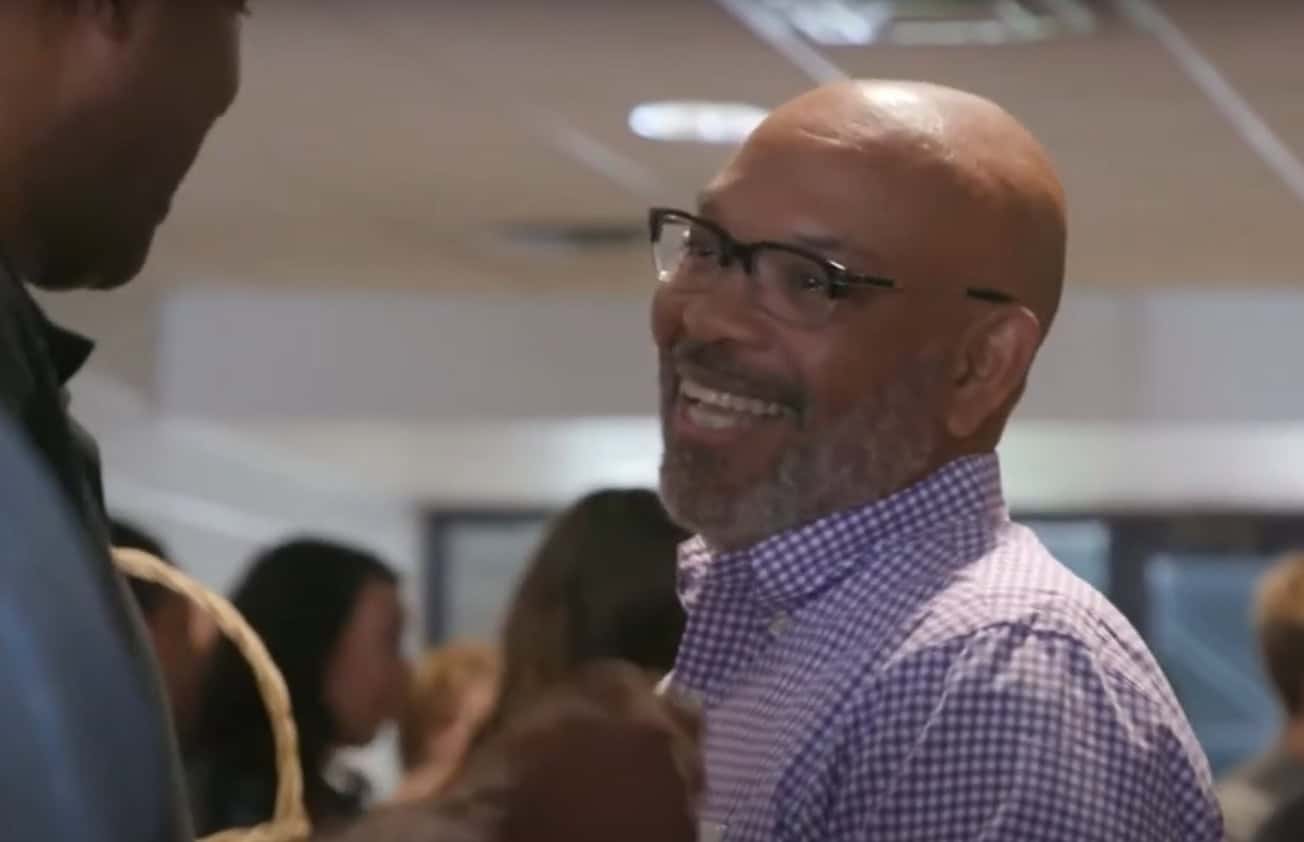 A man with glasses and a beard smiling while talking to another person indoors.